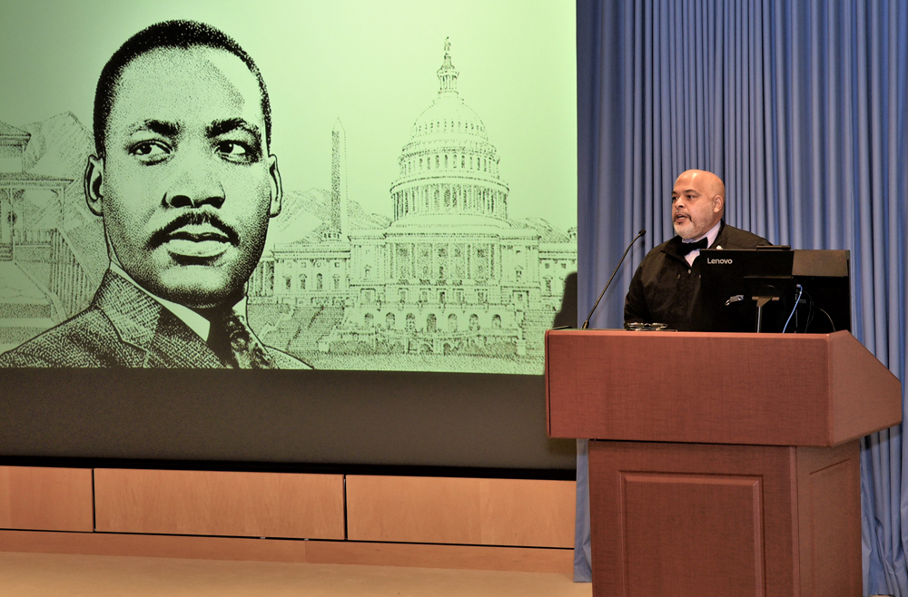 Reverend Brian Dunlop stands behind a podium at the Pennsylvania Hospital Zubrow Auditorium stage with a screen depicting a photo of Martin Luther King, Jr. on the wall behind him
